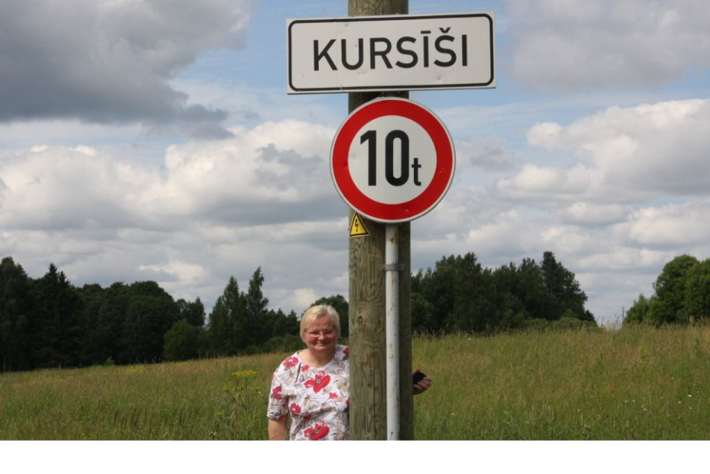 Sieviete ar gaišiem matiem, tērpusies baltā blūzē ar sarkaniem ziediem, stāv pie ceļazīmes lauku ainavā. Uz staba redzama vietas nosaukuma zīme “Kursīši” un zem tās apaļa ceļazīme ar masas ierobežojumu “10 t”. Fonā – zaļi lauki un koki zem mākoņainām debesīm.