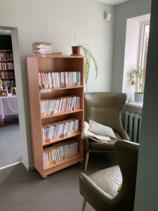 Attēlā ir neliels koka grāmatu plaukts ar baltām grāmatām, blakus tam — zaļi mīkstie atpūtas krēsli un sega. Pie plaukta augšas novietoti daži žurnāli un telpaugs. Telpa atrodas pie loga, pa kuru ienāk dienasgaisma.