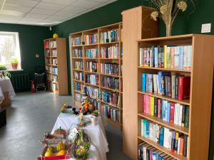 Gar sienu rindojas vairāki augsti, ar grāmatām pilni grāmatu plaukti. Telpa ir gaiša, ar zaļām sienām un dienasgaismu. Priekšplānā redzami galdi ar dekoratīvi sarīkotu izstādi – grozi, augļi un amatniecības izstrādājumi.