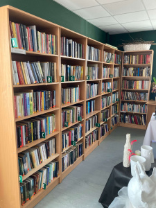 Grāmatu plauktu rinda ar alfabētiskām marķējuma zīmēm uz katras sekcijas (A, B, C u.c.). Plaukti ir piepildīti ar dažādu žanru grāmatām. Priekšplānā – izstādītas dekoratīvas vāzes uz melna auduma. 