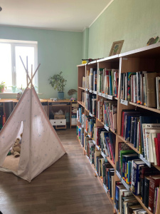 Gara, šaurāka telpa ar grāmatu plauktiem vienā pusē un bērnu tipī telti otrā. Uz grīdas tipī iekšā redzami mīkstie rotaļu lācīši. Fonā — liels logs ar augiem uz palodzes. Telpa ir gaiša un mājīga.